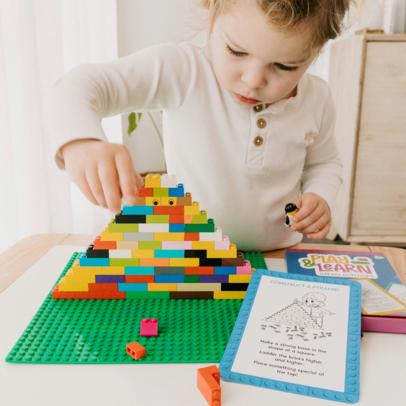 Child building a tall colourful pyramid from bricks, guided by an activity card